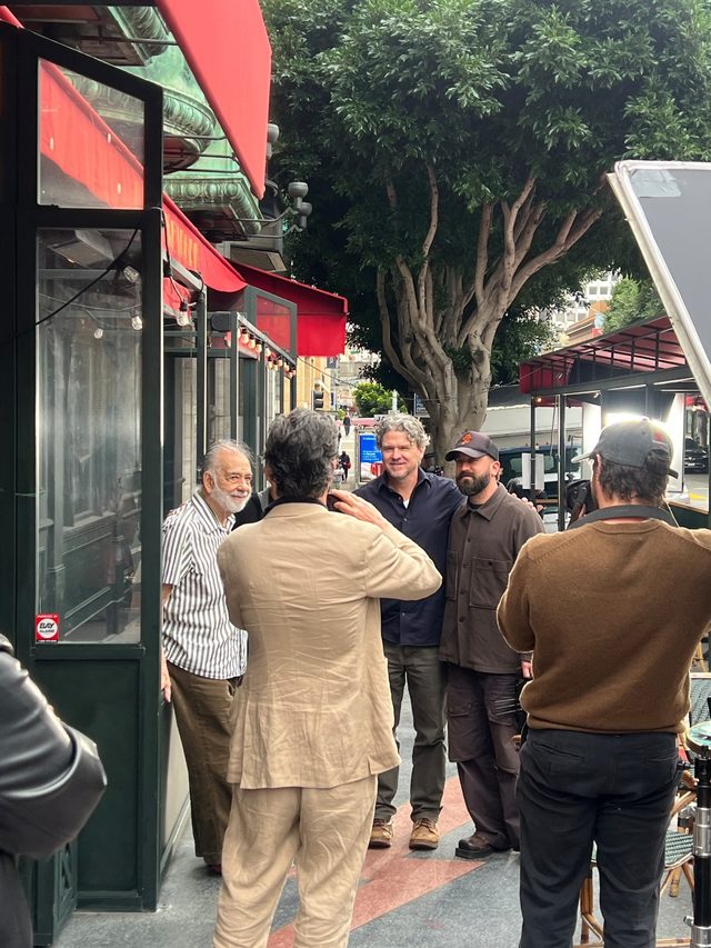 Five men stand on a sidewalk outside a restaurant with red awnings, talking and posing near a large tree, with some photo or film equipment nearby.